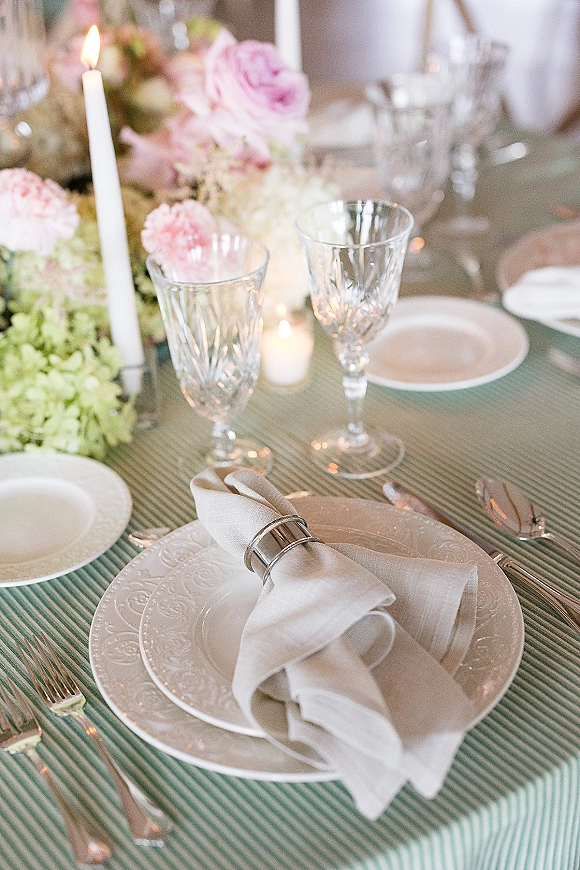Reception tablescape with wedding place setting, crystal wine glasses, white taper candles, and pink rose centerpiece on a striped tablecloth