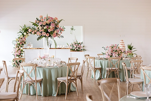 Reception tablescape with round reception tables dressed in mint green table linens, pink rose centerpieces, candles, and cross-back chairs indoors
