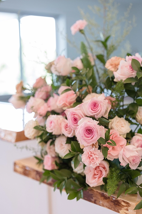 Wedding floral arrangement with pink rose centerpiece of blush and white roses, carnations, and greenery on a wooden ledge by window light