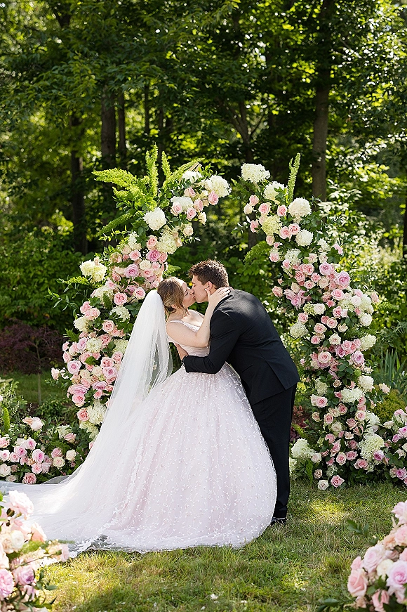 Wedding kiss portrait of the bride and groom kissing under a floral arch with pink roses and hydrangeas on a garden lawn