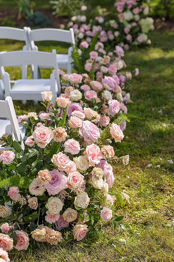 Ceremony aisle flowers with wedding aisle flowers of pink roses and blush blooms, bordered by greenery beside white folding chairs on a lawn