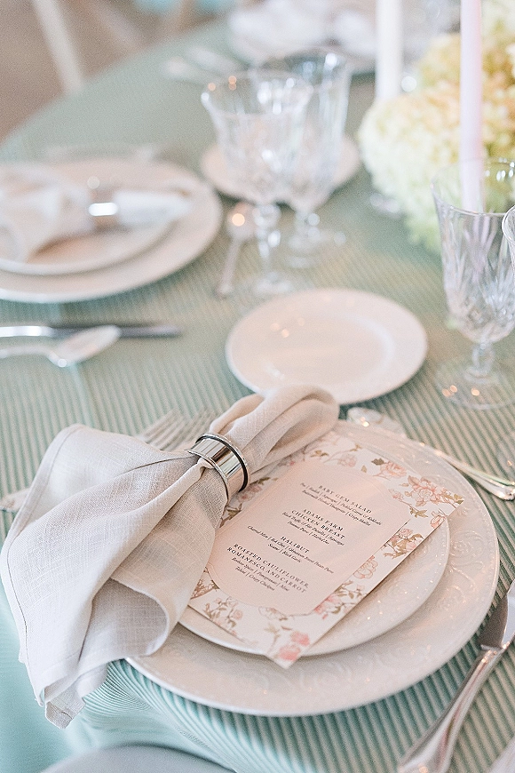 Wedding place setting with layered plates and wedding tablescape menu card on a striped tablecloth, crystal glassware and soft floral centerpiece in bokeh