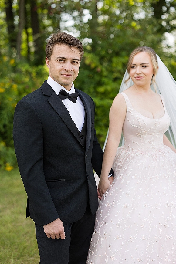 Couple portrait of bride and groom portrait holding hands, bride in lace gown and veil beside groom in black tuxedo on garden lawn