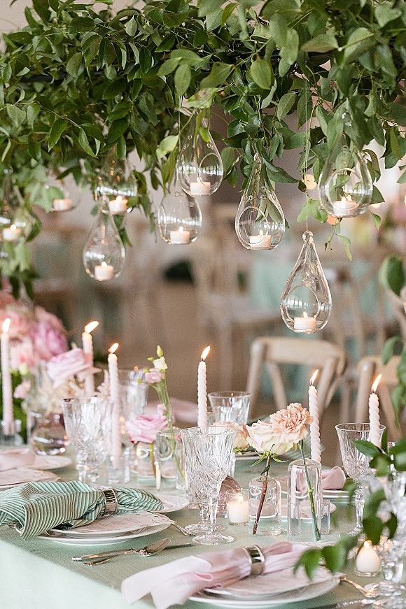 Reception tablescape with wedding table centerpiece of blush roses and greenery garland, crystal goblets, and hanging glass candle holders in a blurred room