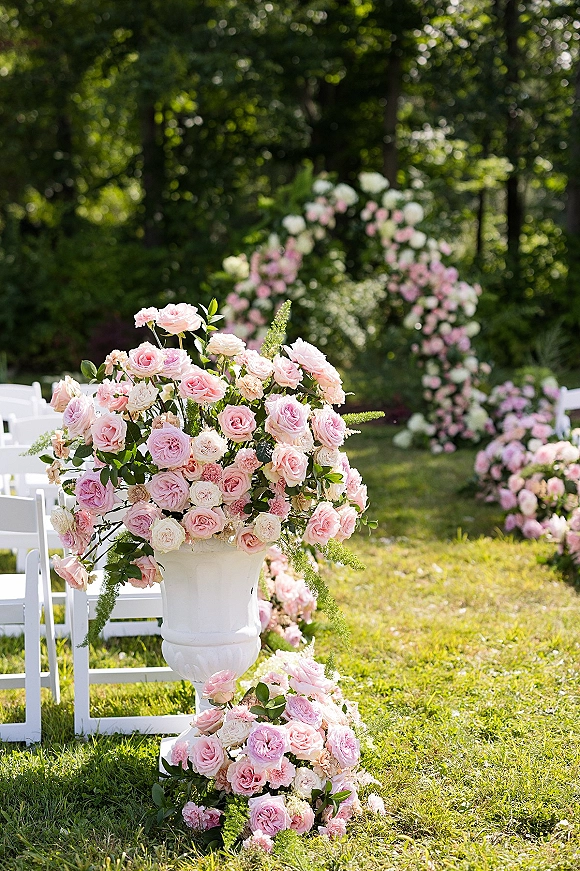 Ceremony aisle decor with outdoor wedding ceremony setup, pink rose and blush florals on white urns leading to a round arch on a garden lawn