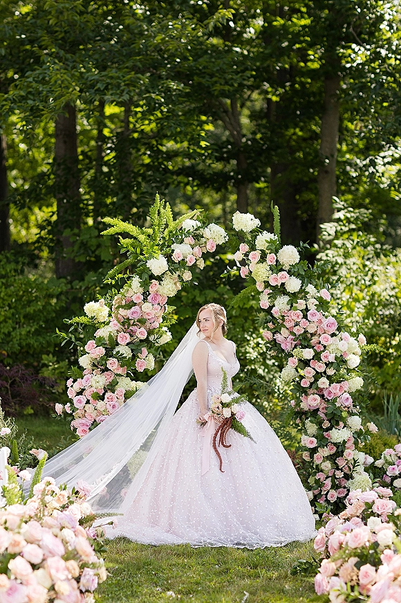 Bridal portrait of a bride in a ball gown wedding dress with long veil, holding a bouquet before a round floral arch in a garden setting