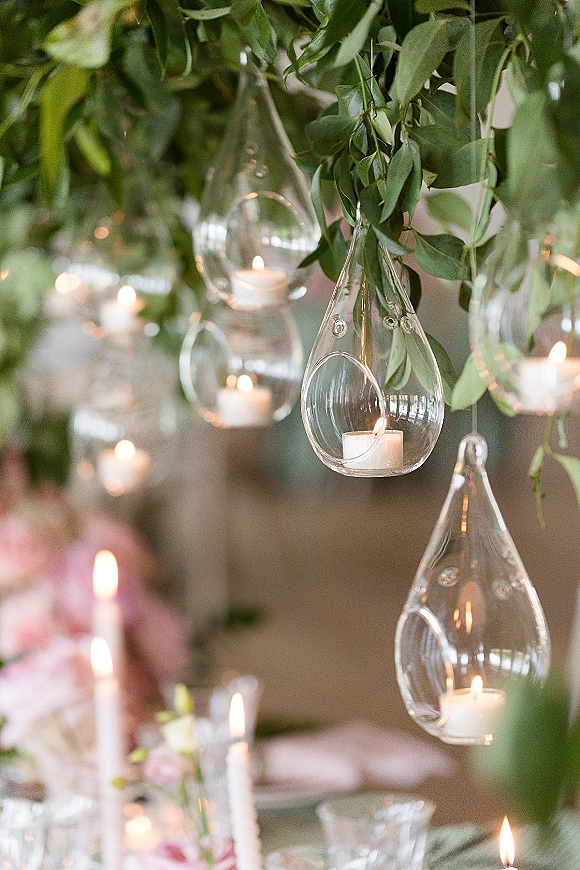 Hanging candle decor with glass hanging votives, glowing tea lights, and greenery garland with pink flowers above a reception table indoors