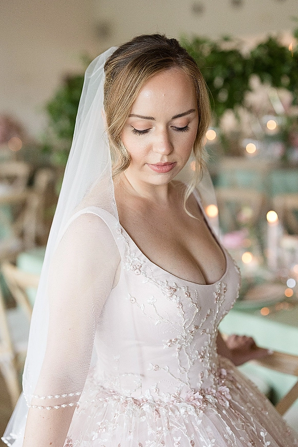 Bridal portrait of bride looking down in a lace dress and wedding veil, pearl-trimmed sheer sleeves, candlelit reception tables behind