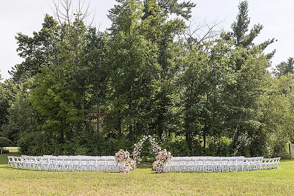 Ceremony setup with outdoor wedding ceremony seating in white folding chairs facing a floral arch with pink flowers on a forest lawn under sky