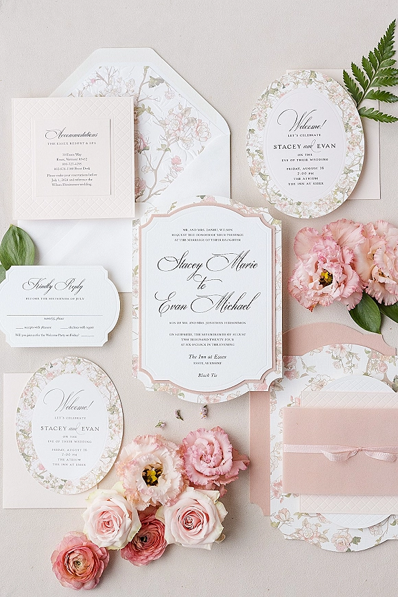 Wedding invitation suite with floral wedding invitations, envelopes and RSVP cards, tied with blush ribbon and framed by pink flowers on a neutral surface