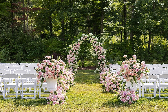 Ceremony setup for an outdoor wedding ceremony with a floral arch, pink and white rose urns, and white chairs lining a grassy aisle among trees