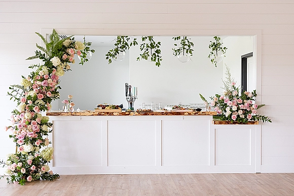 Wedding bar decor with a wedding bar floral garland of roses, hydrangeas, and greenery on a rustic wood bar, with glass orbs above