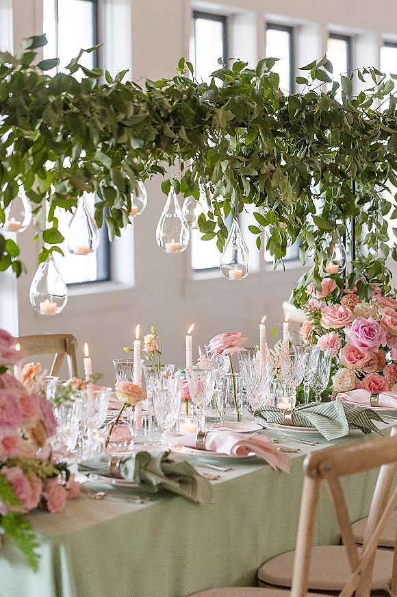 Reception tablescape with a wedding table centerpiece of pink roses, greenery garland, hanging glass votives, and taper candles by large windows