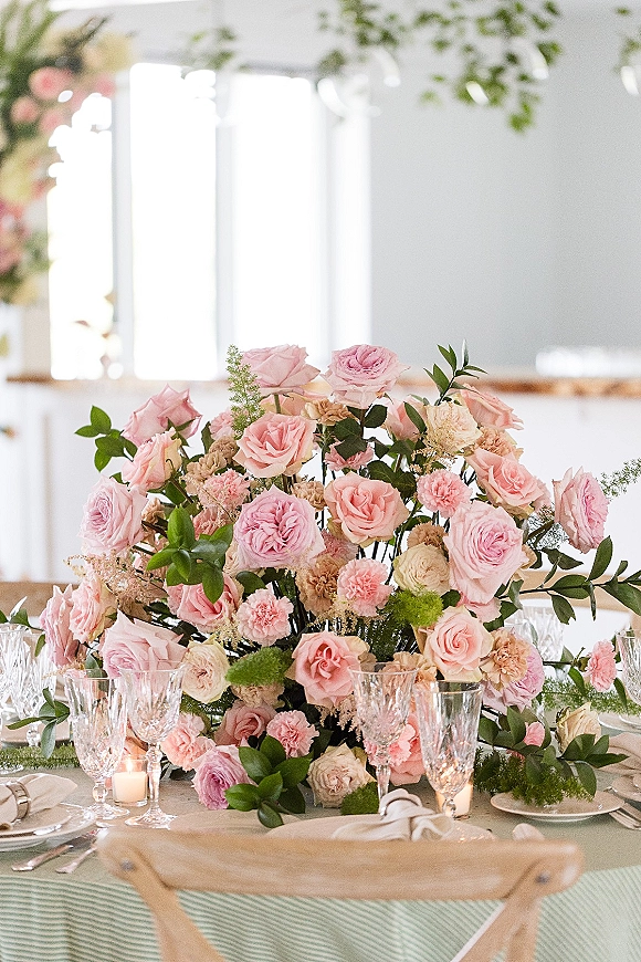 Reception tablescape with pink rose centerpiece, greenery garland, crystal goblets, votive candles, and white plates by bright windows