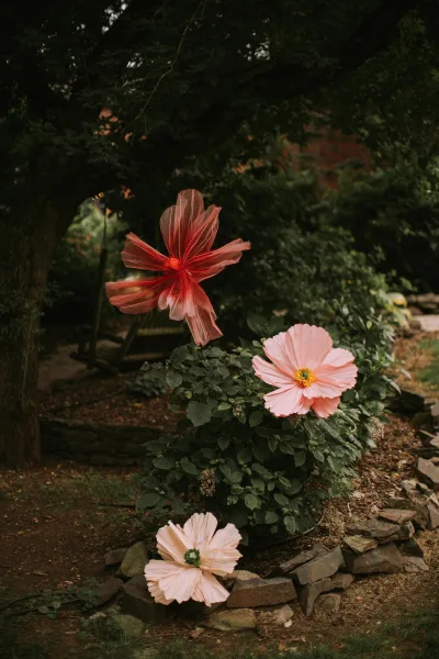 Garden wedding flowers with oversized paper flowers and greenery lining a garden path, framed by shrubs, trees, and a stone border