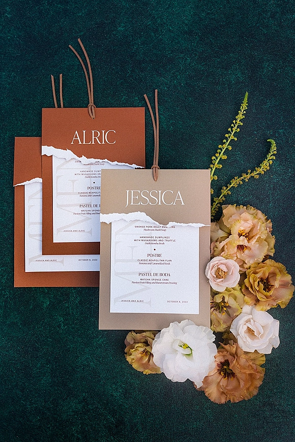 Wedding invitation suite with torn edge paper and layered cardstock tied with leather cord, styled with florals on dark teal surface