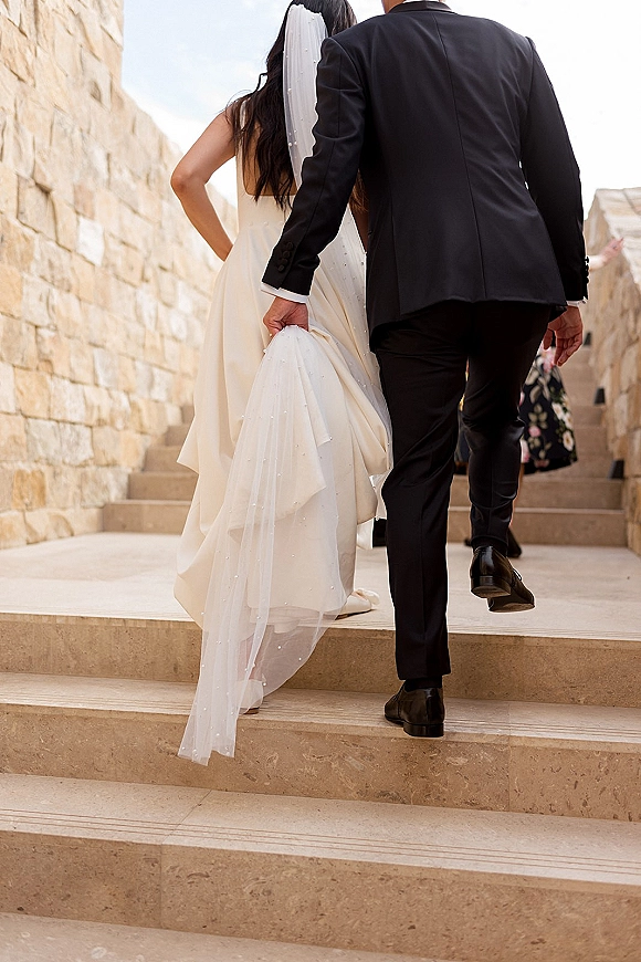 Wedding couple portrait of newlyweds walking away up a stone staircase, bride lifting her satin train beneath a cathedral veil as guests look on
