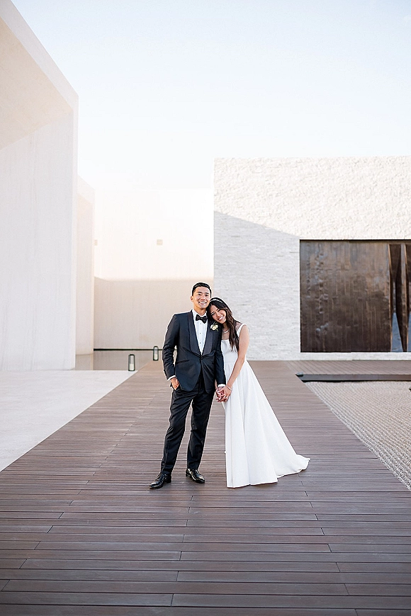 Couple portrait of bride in a minimalist gown and groom in black tuxedo holding hands on a wooden deck by white modern walls