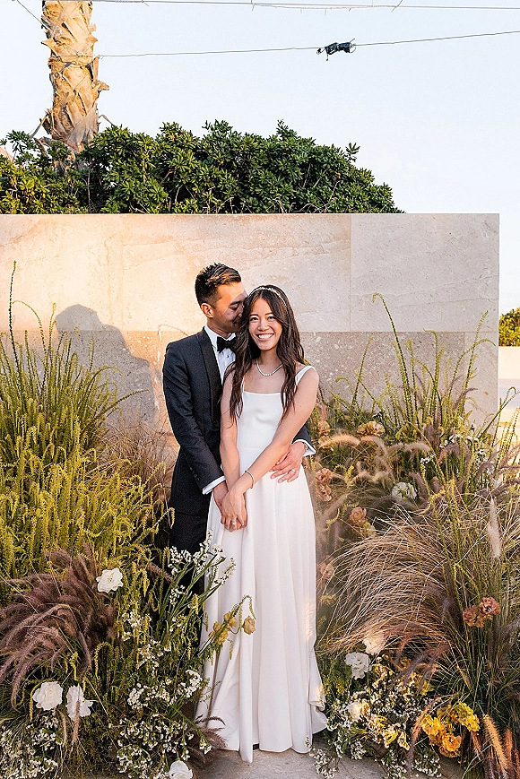 Couple portrait of bride and groom hug as he kisses her cheek, bride in satin dress and pearls by stone wall and string lights