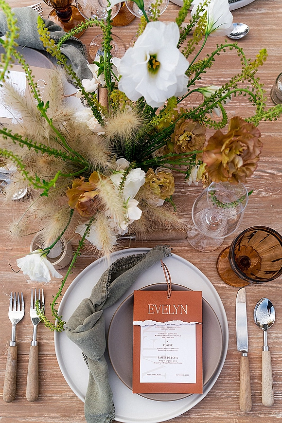 Reception tablescape with wedding table centerpiece of pampas grass and white blooms, amber goblets, taper candle, and menus on a wood table