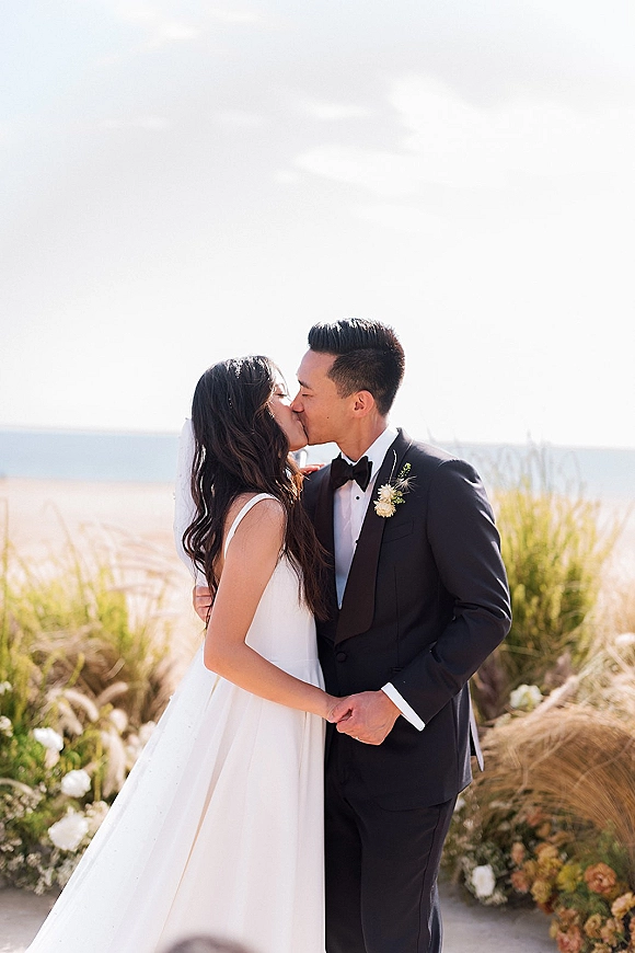 Wedding kiss portrait of bride and groom kissing in a close embrace on a beach, her veil blowing by ocean dunes and white florals