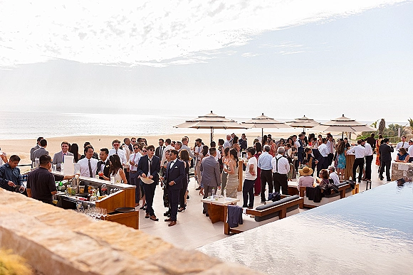 Wedding cocktail hour with guests in suits and dresses mingling at an outdoor bar by a pool, ocean backdrop, and patio umbrellas