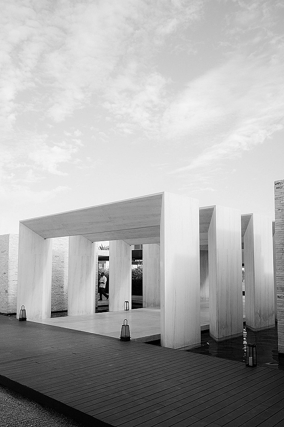 Modern ceremony venue with lanterns lining a wood deck beside a water feature, framed by minimalist architecture under a cloudy sky