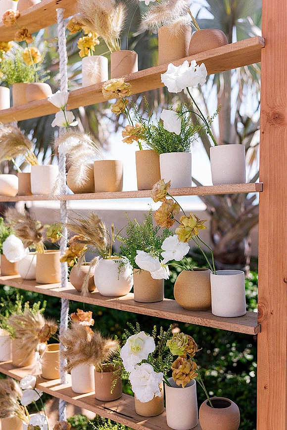 Wedding floral backdrop with ceremony backdrop flowers on wood shelves, ceramic bud vases and pampas grass, set outdoors by palm trees