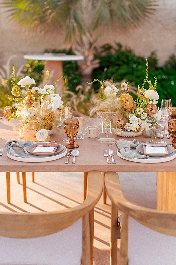 Reception tablescape with pampas grass centerpiece, amber goblets, menus and place cards on a wood table set on a stucco patio outdoors