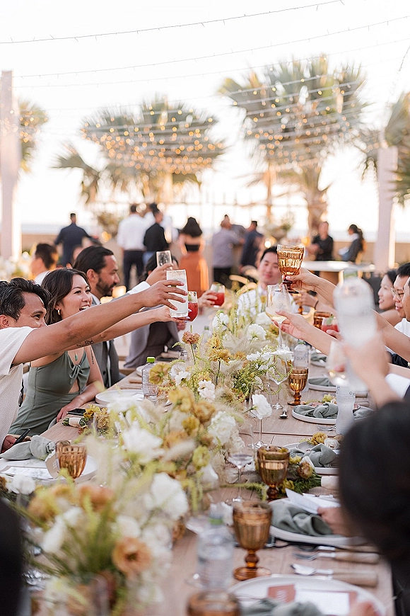 Wedding reception toast with guests raising amber goblets over a long wooden table, string lights glowing on a coastal patio with palms