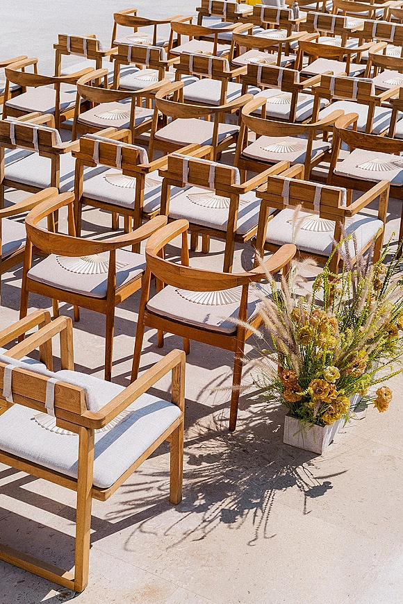 Ceremony seating with wedding chair layout of wood chairs, white cushions and fans beside a floral planter on a sunlit stone patio
