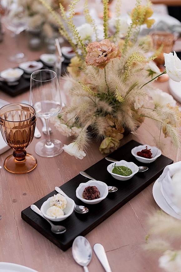 Reception tablescape with wedding table centerpiece of pampas grass and white flowers, amber goblets and shared wooden serving board on wood table