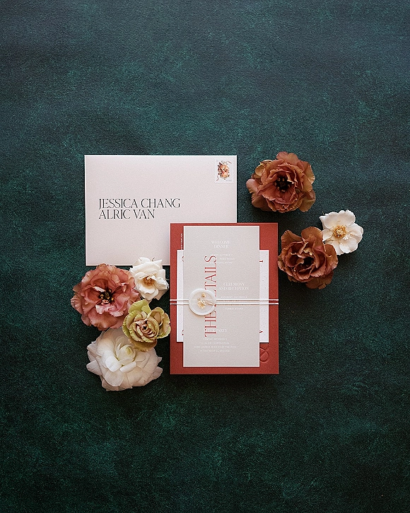 Wedding invitation suite flat lay with envelopes, RSVP card, belly band and wax seal, styled with flowers on a dark green surface