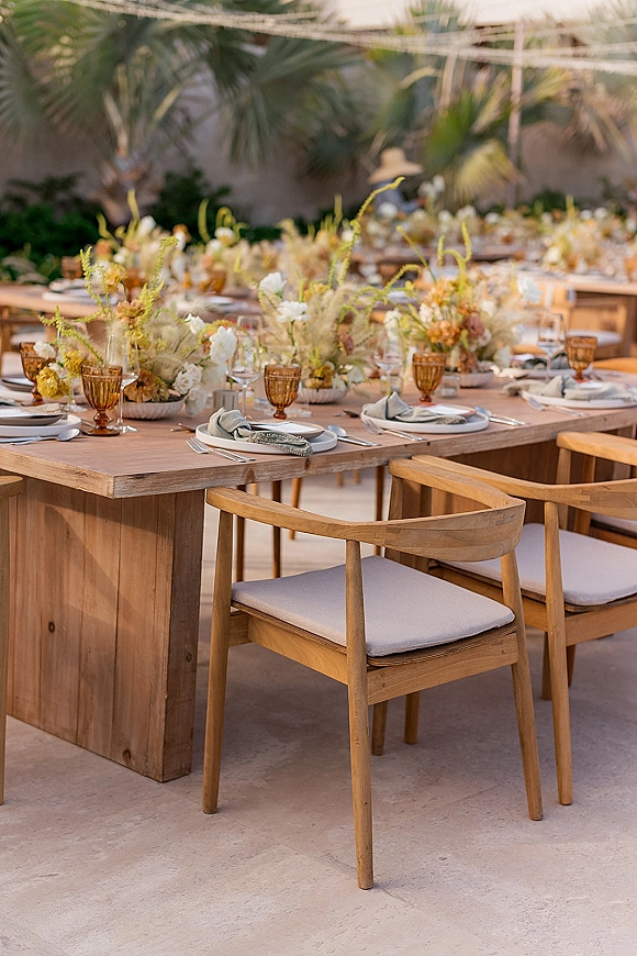 Reception tablescape with amber goblets, white plates, floral centerpieces, and taper candles on a long wooden table on a palm-lined patio