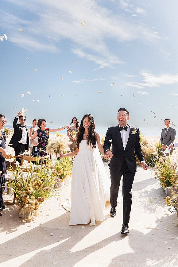 Wedding recessional as bride and groom walk the aisle on a beach, guests toss flower petals under blue sky and ocean horizon