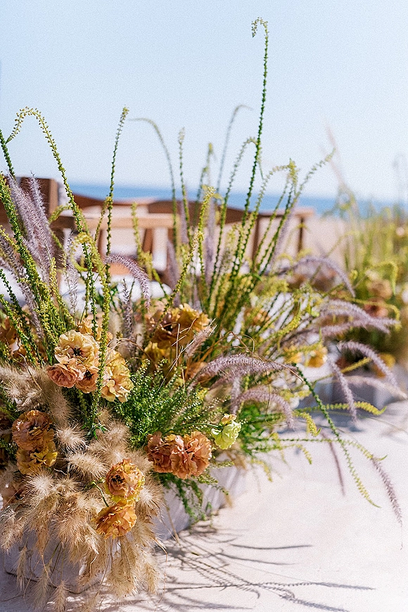 Wedding centerpiece with peach flowers and dried grasses in a vase on an outdoor table, wooden chairs and ocean view behind