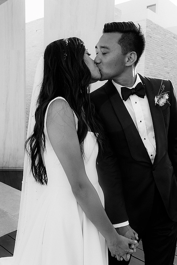 Wedding kiss portrait of bride and groom kissing, holding hands, with a flowing veil beside modern columns and a brick wall walkway