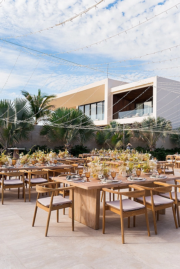 Outdoor reception tablescape with wood banquet tables wedding details, amber goblets, floral centerpieces, and string lights in a palm courtyard