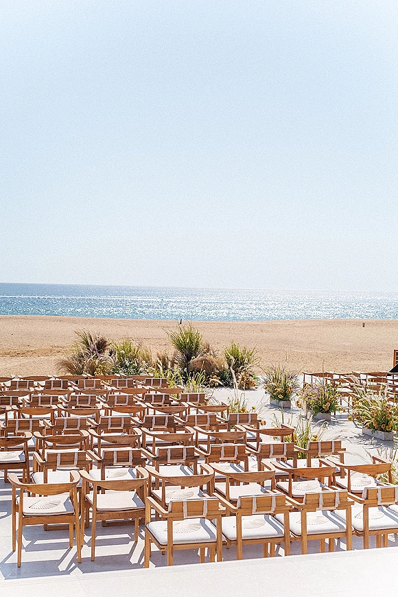 Beach ceremony setup with wood chairs and a white aisle runner lined with floral greenery, pampas accents, facing the ocean horizon