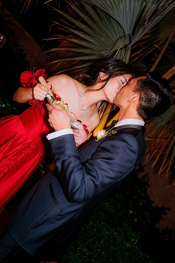 Wedding kiss as groom dips bride in a red bridal gown, sharing a dessert bite with spoon under palm leaves at night