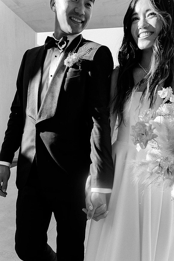 Couple portrait in black and white wedding portrait style, bride and groom holding hands, smiling indoors as she carries a bouquet