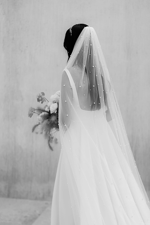 Bridal portrait of a back view bride in a pearl wedding veil and sleek gown, holding a bouquet against a plain wall background