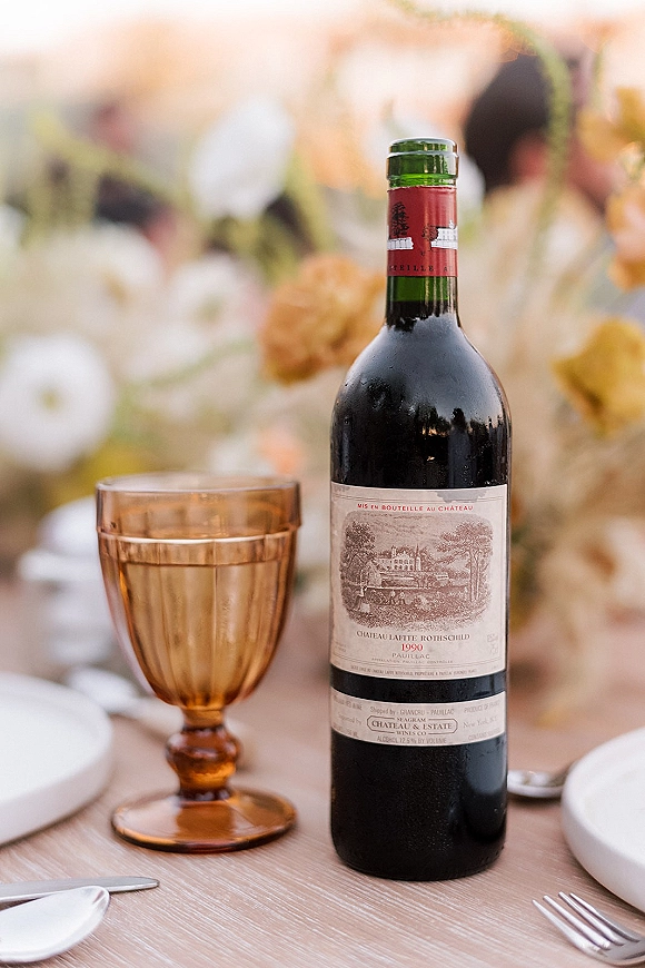 Wedding wine bottle on a reception table with amber goblet, place setting, and floral centerpiece, with guests blurred in the background