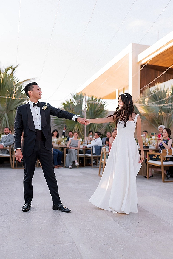 First dance outdoors as bride in a simple wedding dress twirls with groom in black tuxedo under string lights on a patio with palm trees