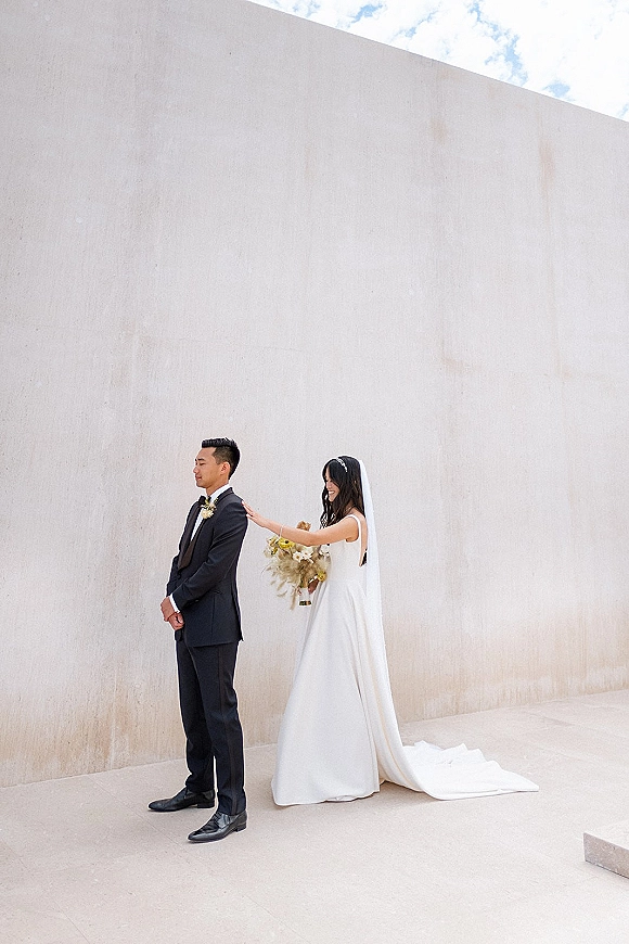 First look moment as bride taps groom’s shoulder, holding a bouquet, her veil trailing beside a white wall under cloudy sky