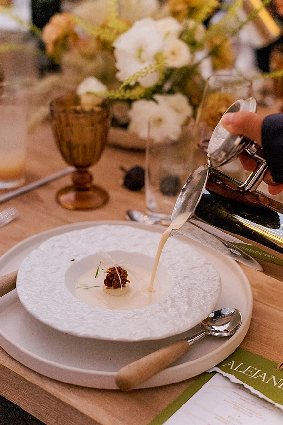 Wedding dinner course with soup in a white patterned bowl on a plate, set with wood-handled flatware, amber goblet, and menu card on a wood table
