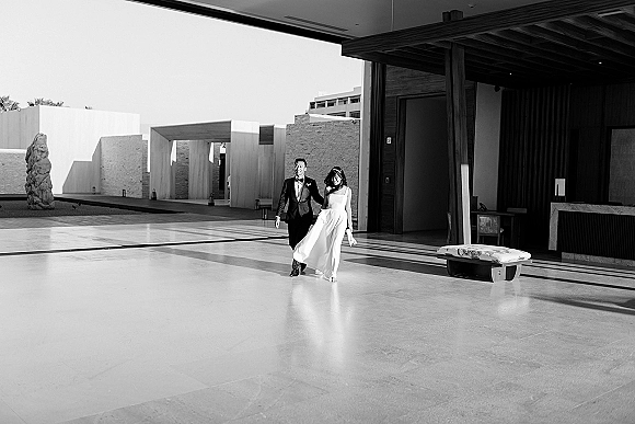 Couple portrait of bride with veil and groom in tuxedo walking hand in hand across a modern courtyard patio with bold shadows