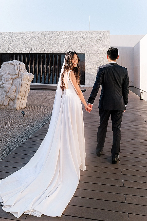Couple portrait of bride and groom holding hands, walking away in a courtyard by modern facade, bride looking back with veil train