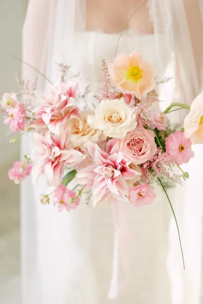 Bridal bouquet of pastel bridal bouquet roses, dahlias, cosmos, and greenery with ribbon, held against a wedding dress and veil