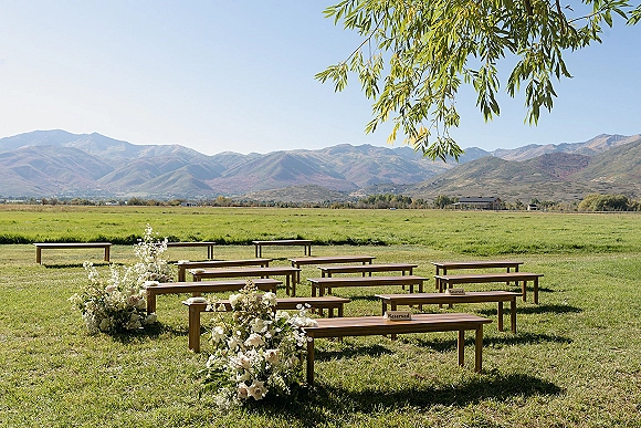 Ceremony setup with rustic wood benches and white floral greenery accents lining the aisle in a mountain meadow under blue sky